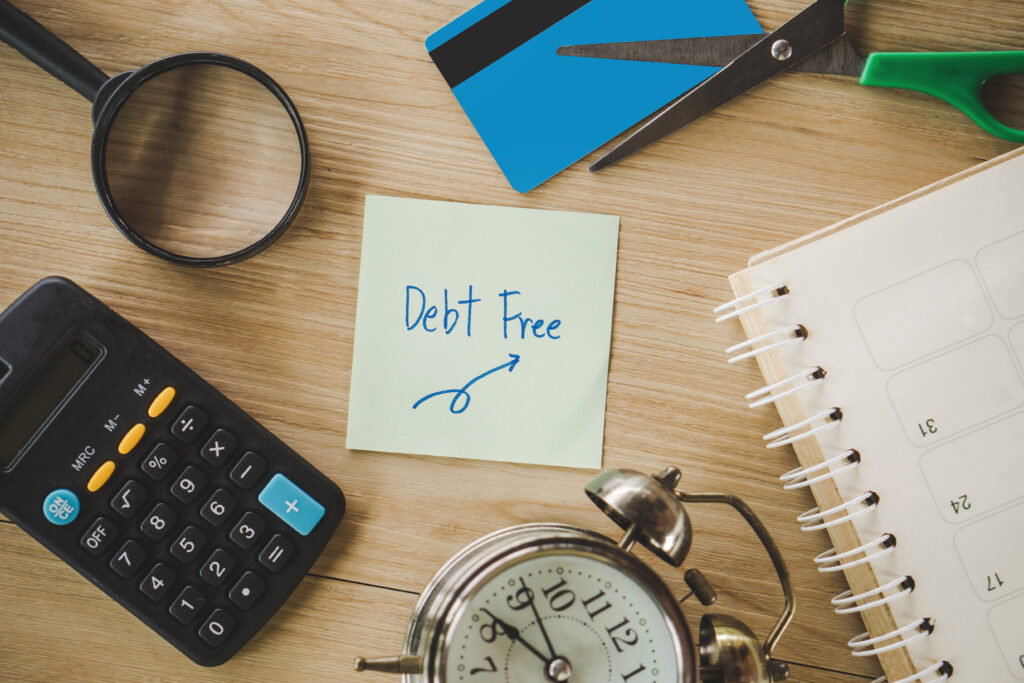 Sticky note with “Debt Free” written on it surrounded by a calculator, scissors, a cut credit card, a planner, and an alarm clock.