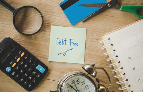 Sticky note with “Debt Free” written on it surrounded by a calculator, scissors, a cut credit card, a planner, and an alarm clock.