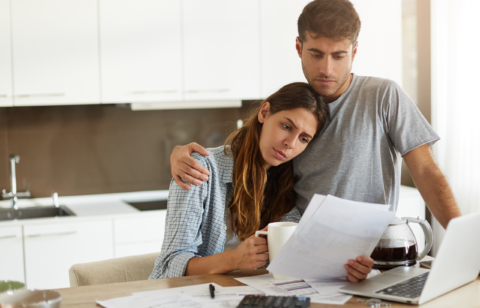 A worried couple sits at a kitchen table reviewing bills and financial documents, with a laptop, coffee pot, and calculator in front of them.