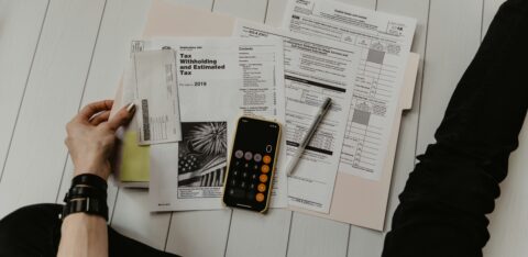 Person sitting on the floor with tax documents, calculator app open on a smartphone.