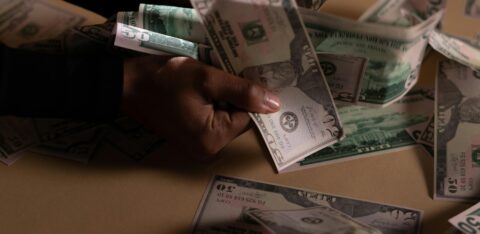 Person counting a large stack of U.S. dollar bills on a beige table under dramatic lighting.