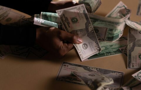 Person counting a large stack of U.S. dollar bills on a beige table under dramatic lighting.