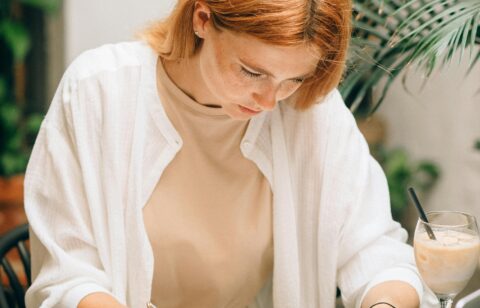Woman focused on filling out financial documents at a desk with a laptop, calculator, magnifying glass, and iced coffee.