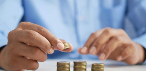 Close-up of a person in a blue shirt stacking coins into small piles on a table, representing saving or budgeting money.