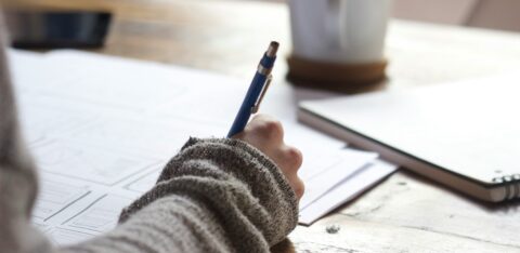 Close-up of a person writing on paper at a wooden desk with a notebook and coffee mug nearby.