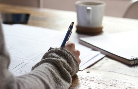Close-up of a person writing on paper at a wooden desk with a notebook and coffee mug nearby.