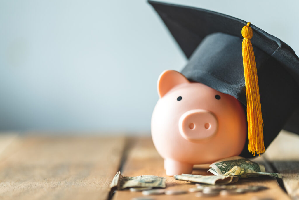 Pink piggy bank wearing a black graduation cap, surrounded by scattered dollar bills and coins on a wooden surface.