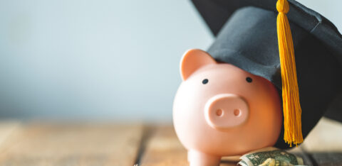 Pink piggy bank wearing a black graduation cap, surrounded by scattered dollar bills and coins on a wooden surface.