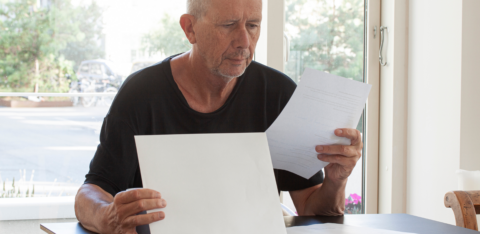 Older man sitting at a table reading documents in a brightly lit room.