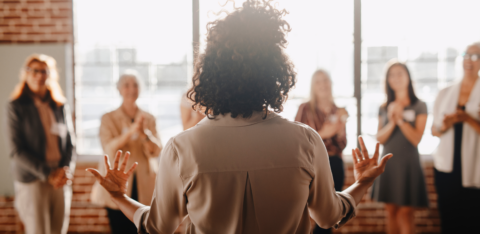Back view of a woman speaking to a group of people in a bright room, with several women standing and clapping.