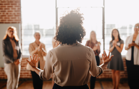 Back view of a woman speaking to a group of people in a bright room, with several women standing and clapping.