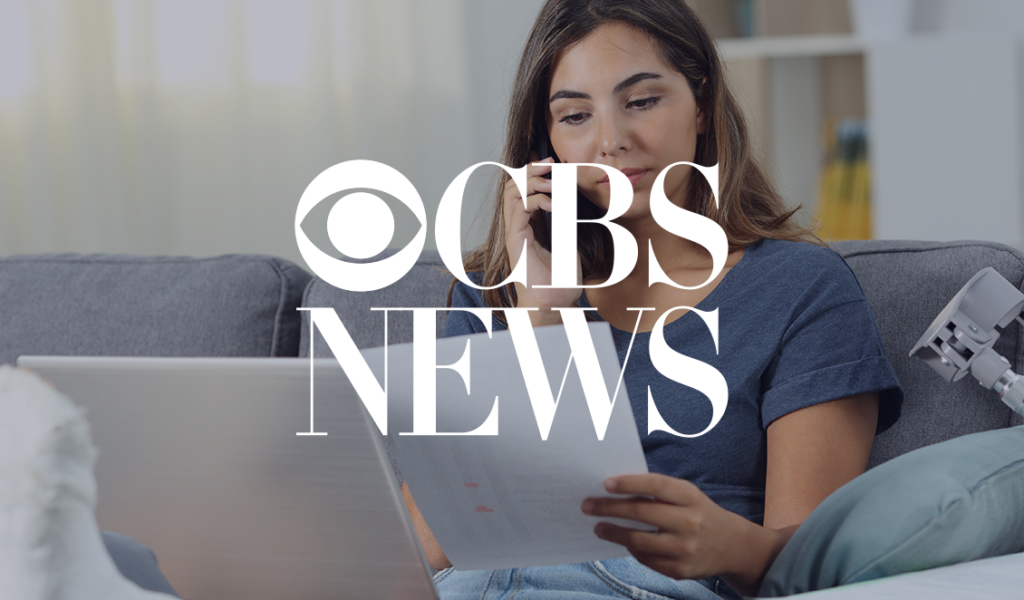 Young woman sitting on a couch looking at paperwork and speaking on the phone, with CBS News logo overlaid.