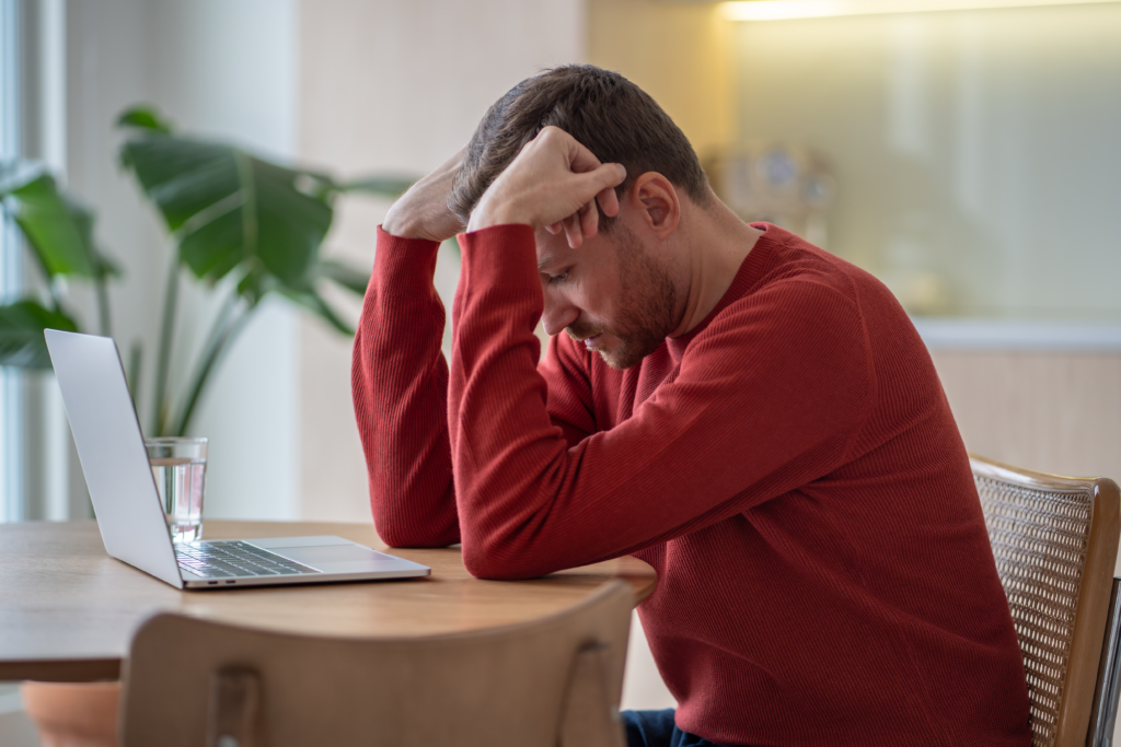 Man in a red sweater sitting at a table with his head in his hands, looking stressed while using a laptop.