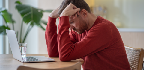 Man in a red sweater sitting at a table with his head in his hands, looking stressed while using a laptop.