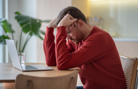 Man in a red sweater sitting at a table with his head in his hands, looking stressed while using a laptop.
