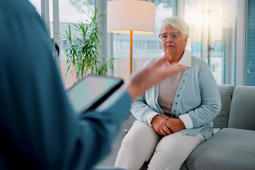 Older woman sitting on a couch listening to a professional with a tablet during a counseling session.