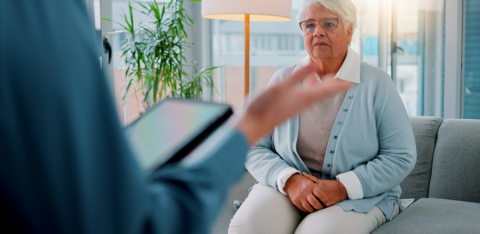 Older woman sitting on a couch listening to a professional with a tablet during a counseling session.