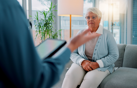 Older woman sitting on a couch listening to a professional with a tablet during a counseling session.