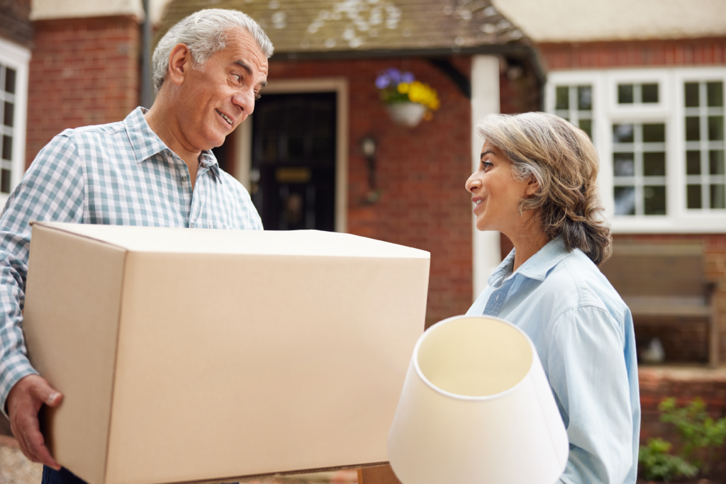 Older couple smiling at each other while carrying moving boxes and a lamp outside a brick home.