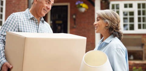 Older couple smiling at each other while carrying moving boxes and a lamp outside a brick home.