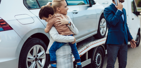 Woman holding a child beside a car being towed, while a man in a suit makes a phone call.