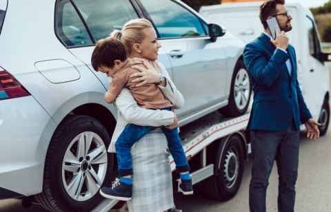 Woman holding a child beside a car being towed, while a man in a suit makes a phone call.
