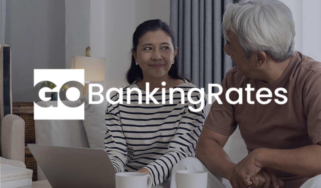 A senior couple sits on a couch talking, with a laptop and coffee mugs in front of them. The GoBankingRates logo is overlaid on the image.