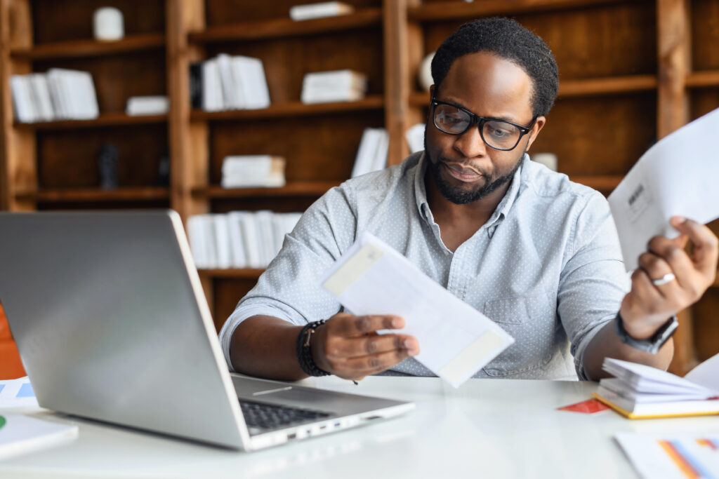 Man wearing glasses sitting at a desk, reviewing mail and bills while using a laptop.