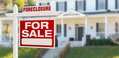 Red "For Sale" sign with "Foreclosure" label in front of a large, white house.