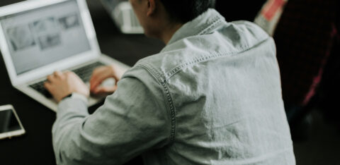 Person working on a laptop at a desk, viewed from behind, with a smartphone placed nearby.