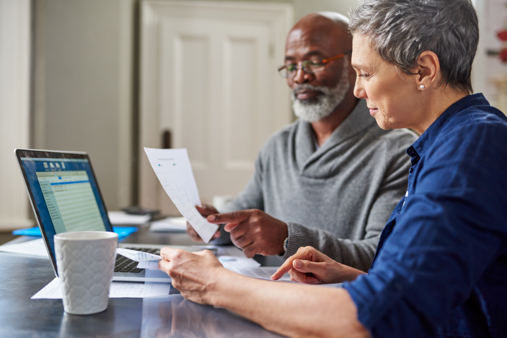Older couple reviewing documents together at a kitchen table, with a laptop open and papers spread out in front of them.
