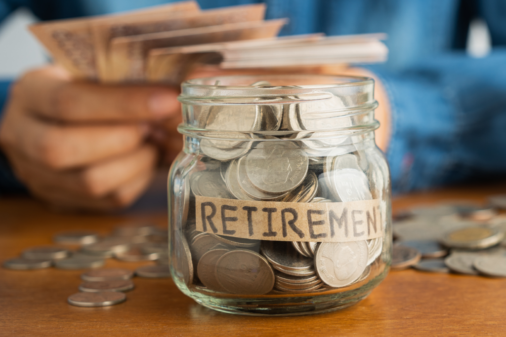 Glass jar labeled 'Retirement' filled with coins, with a person in the background holding currency notes.