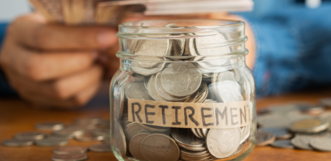 Glass jar labeled 'Retirement' filled with coins, with a person in the background holding currency notes.