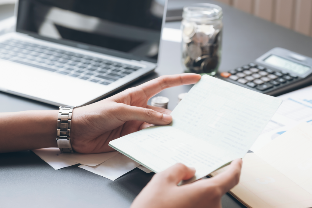 Hands holding a financial statement or passbook at a desk with budgeting tools, including a laptop, calculator, coins, and paperwork.