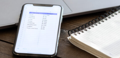 Smartphone displaying a personal monthly budget spreadsheet next to a spiral notebook and laptop on a wooden desk.