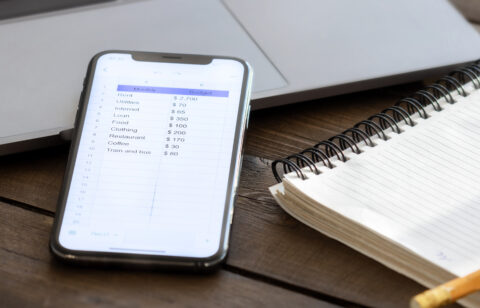 Smartphone displaying a personal monthly budget spreadsheet next to a spiral notebook and laptop on a wooden desk.