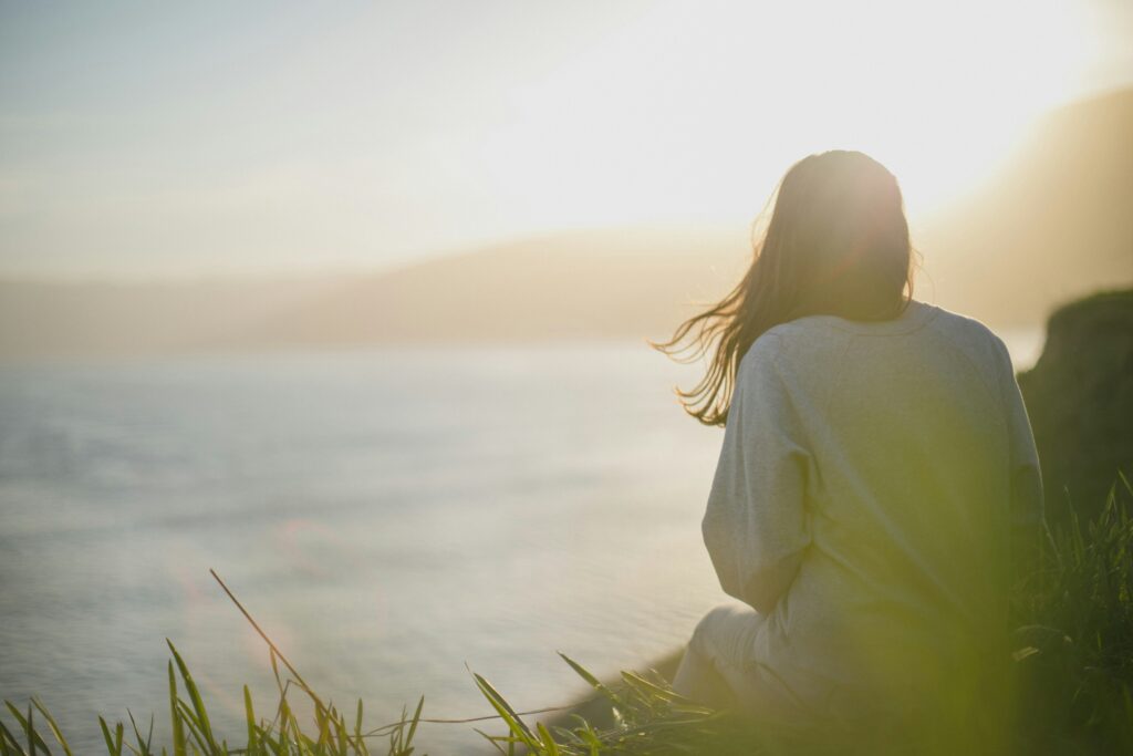 Person sitting alone near a body of water at sunset, gazing into the distance in quiet reflection.