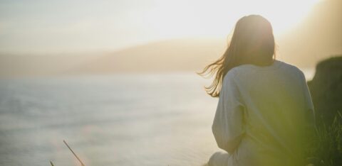 Person sitting alone near a body of water at sunset, gazing into the distance in quiet reflection.