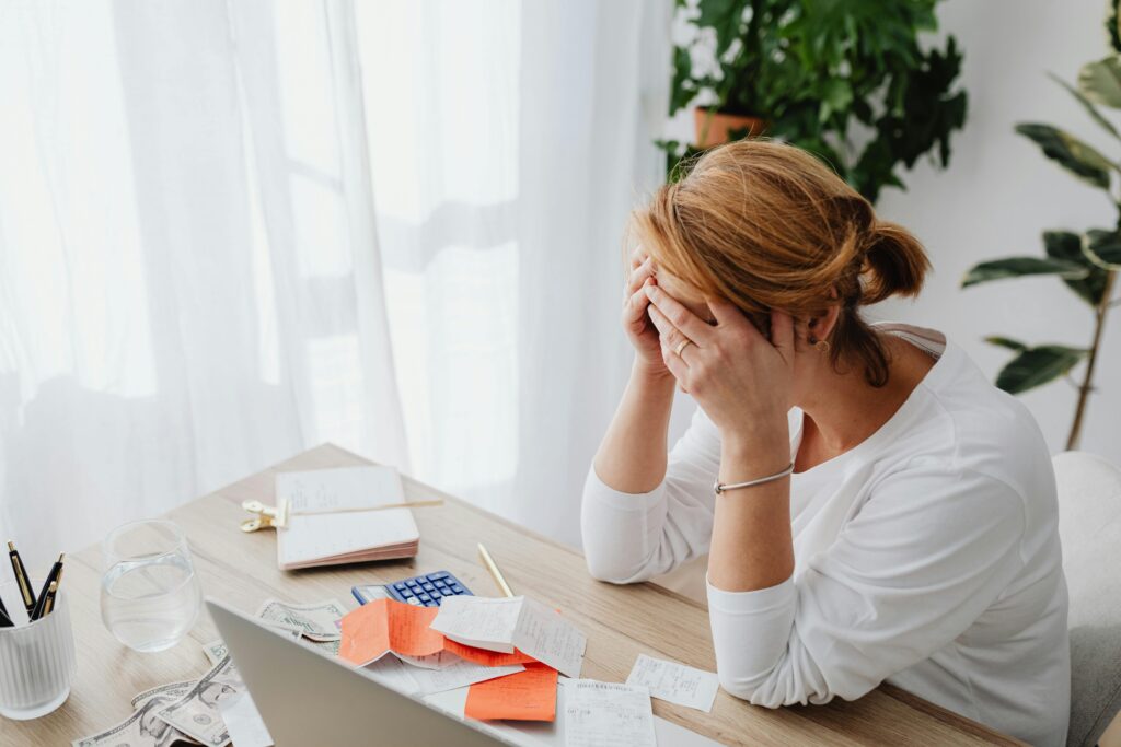 Woman sitting at a desk with her head in her hands, surrounded by bills and receipts, appearing stressed.