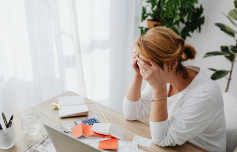 Woman sitting at a desk with her head in her hands, surrounded by bills and receipts, appearing stressed.