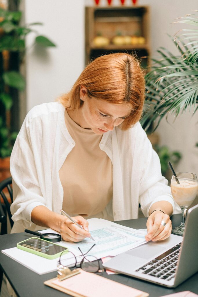 Woman reviewing documents and writing on paper at a desk with a laptop, calculator, and iced coffee