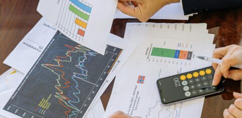 Two professionals review printed charts and graphs at a meeting table, using a calculator and laptop to analyze financial or marketing data.