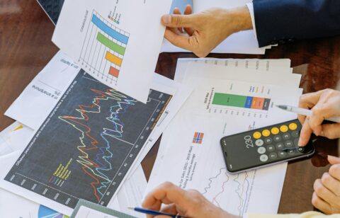 Two professionals review printed charts and graphs at a meeting table, using a calculator and laptop to analyze financial or marketing data.