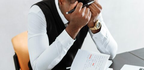Man sitting at a desk with his head in his hands, surrounded by a laptop, notebook, and hourglass.