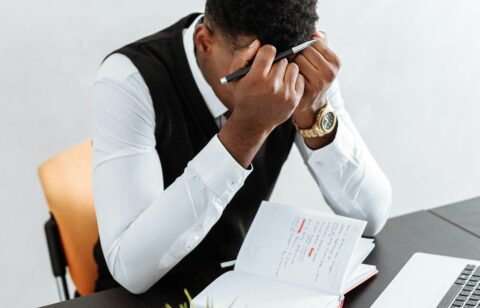 Man sitting at a desk with his head in his hands, surrounded by a laptop, notebook, and hourglass.