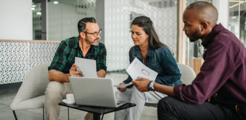 Three adults sit together reviewing documents and charts with a laptop during a discussion in an office.
