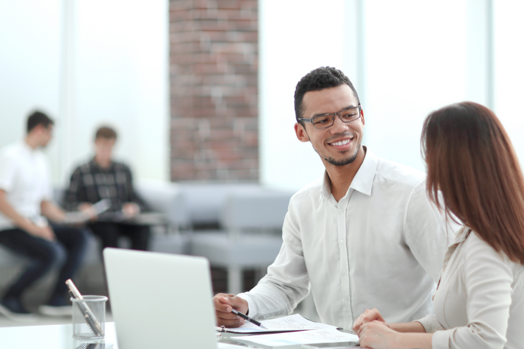 Two professionals having a conversation at a desk with documents and a laptop in a bright office setting.