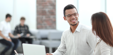 Two professionals having a conversation at a desk with documents and a laptop in a bright office setting.