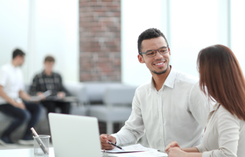 Two professionals having a conversation at a desk with documents and a laptop in a bright office setting.