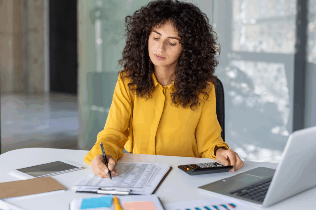 Woman in a yellow blouse using a calculator while reviewing a financial spreadsheet at her desk.